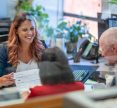 A Hispanic woman sits at her desk at work and smiles while reviewing paperwork with a senior adult couple.